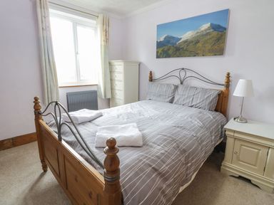 A bedroom with a wooden bed frame, striped bedding, two white towels on the bed, a window with curtains, a chest of drawers, a bedside table with a lamp, and a landscape painting on the wall at Tan Y Fron in Dyffryn Ardudwy