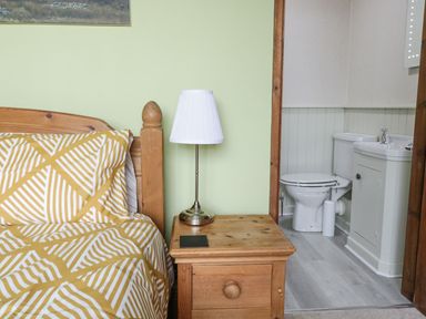 A bedroom with a wooden bed yellow patterned bedding a wooden nightstand with a lamp next to a bathroom with a toilet and sink at Tan Y Fron in Dyffryn Ardudwy