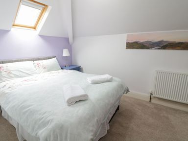 A bedroom with a bed with white and floral bedding a bedside table with a lamp a skylight window and a landscape painting on the wall at Tan Y Fron in Dyffryn Ardudwy