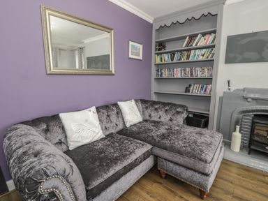 A living room with a purple wall, a gray velvet sofa with white pillows, a mirror, a bookshelf with books and DVDs, and a gray tiled fireplace at Tan Y Fron in Dyffryn Ardudwy