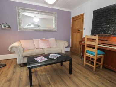A living room with a beige sofa and pink cushions a black coffee table with magazines a wooden chair with a blue cushion a piano and a large mirror on a purple wall at Tan Y Fron in Dyffryn Ardudwy