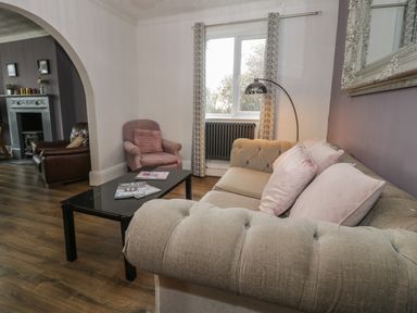 A living room with a beige sofa with pink cushions a black coffee table a pink armchair a window with curtains a floor lamp and an archway leading to another room at Tan Y Fron in Dyffryn Ardudwy