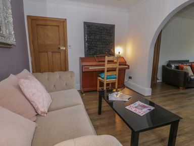 A living room with a beige sofa, wooden door, piano with a chair, black coffee table with magazines, and a brown leather sofa with cushions at Tan Y Fron in Dyffryn Ardudwy