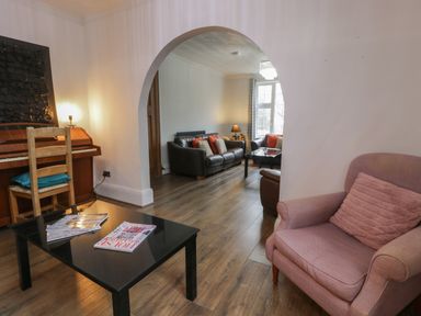 A living room with a black coffee table with magazines a pink armchair a piano with a lamp and a wooden chair and an archway leading to another seating area with a window at Tan Y Fron in Dyffryn Ardudwy
