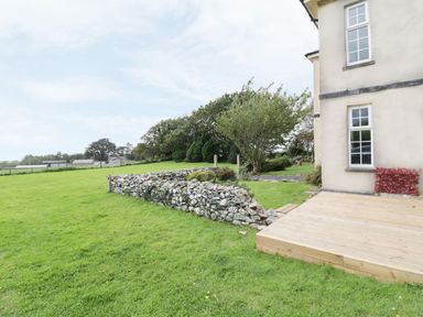 The exterior of a house with a stone wall a wooden deck a grassy lawn and trees at Tan Y Fron in Dyffryn Ardudwy