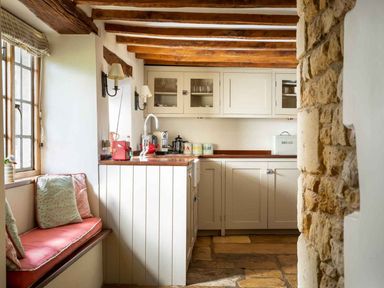 A kitchen with wooden beams stone wall window seat with cushions and cabinets at Hope Cottage in Longborough