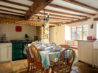 A dining area with a table set with dishes and glasses wooden chairs a window bench with cushions a green stove and wooden ceiling beams at Hope Cottage in Longborough