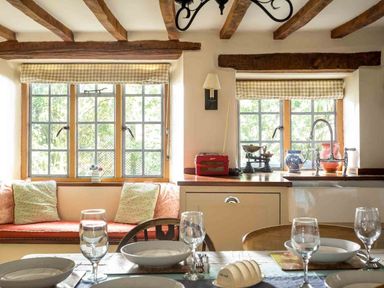 A kitchen and dining area with wooden beams on the ceiling a window seat with cushions and a table set with plates and glasses at Hope Cottage in Longborough