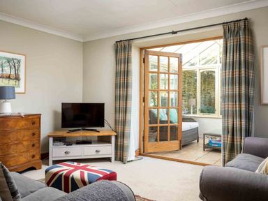 A living room with a television on a stand a wooden chest of drawers and a door leading to a sunroom at Hope Cottage in Longborough