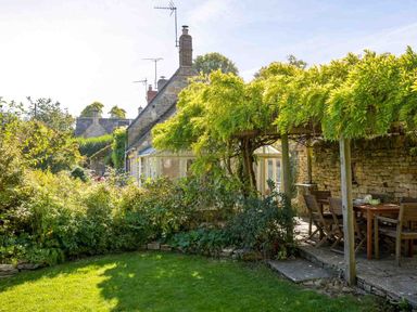 A garden with green plants a covered patio with wooden table and chairs and a stone cottage at Hope Cottage in Longborough
