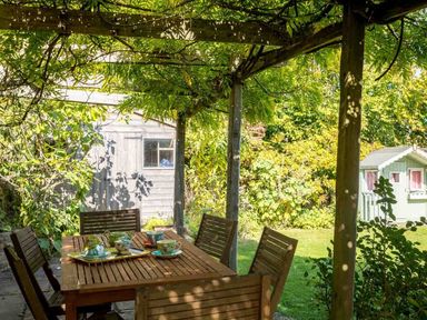 An outdoor wooden table with chairs on a patio under a pergola covered in greenery with a shed and garden in the background at Hope Cottage in Longborough