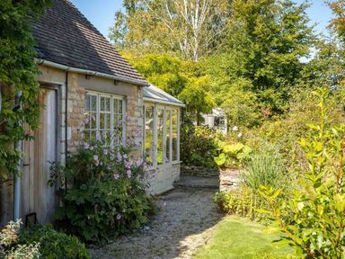 A stone cottage with a wooden door a glass porch and a garden with flowering plants and trees at Hope Cottage in Longborough