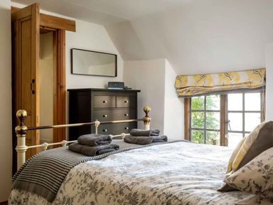 A bedroom with a bed dressed in floral bedding with folded towels on top a black dresser a wooden door and a window with a yellow floral patterned blind at Hope Cottage in Longborough