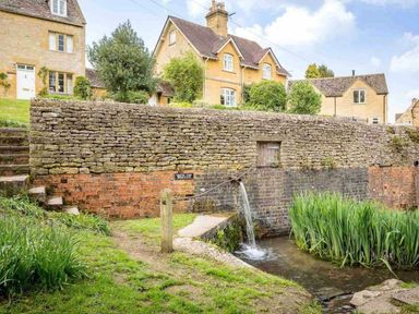 Stone houses behind a stone wall with water flowing from a pipe into a small pond surrounded by grass and plants at Hope Cottage in Longborough
