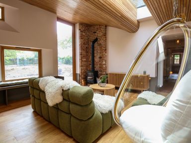 A living room with a green sofa, wood stove with brick wall, round wooden tables, hanging chair, and large windows at Oak Cottage in Somerleyton