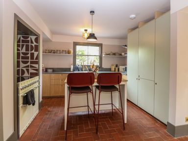 A kitchen with two red chairs at an island and shelves with jars at Oak Cottage in Somerleyton
