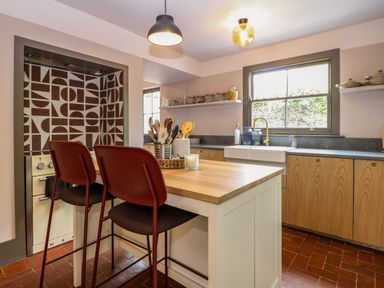 A kitchen with wooden cabinets a white island with two red chairs and patterned tiles behind the stove at Oak Cottage in Somerleyton