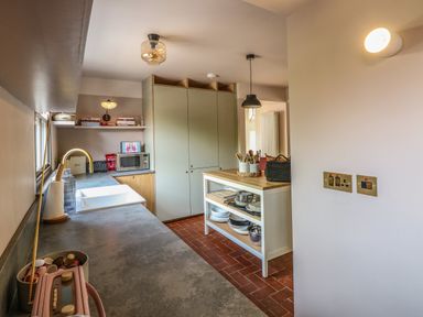 A kitchen with a countertop sink, cabinets, microwave, shelves with books, and a kitchen island with utensils at Oak Cottage in Somerleyton