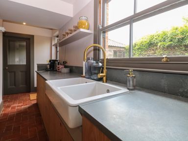 A kitchen countertop with a double sink and golden faucet next to a window with shelves holding jars above at Oak Cottage in Somerleyton