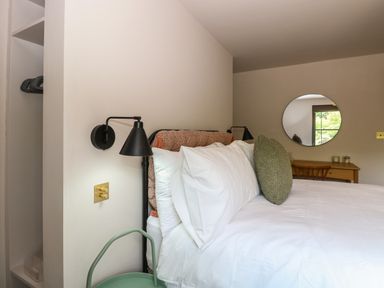 A bedroom with a bed with white sheets green pillow wall lamps and a round mirror above a wooden desk at Oak Cottage in Somerleyton
