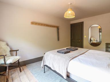 A bedroom with a bed, two folded towels, a wooden chair with a sheepskin, a wall mirror, and a door at Oak Cottage in Somerleyton