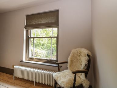 A room with a window with a brown blind a white radiator below and a wooden chair with a white sheepskin throw at Oak Cottage in Somerleyton