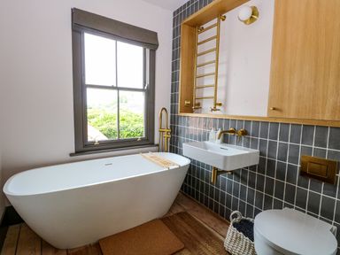 A bathroom with a white freestanding bathtub a window a wall-mounted sink with gold fixtures and a towel rail at Oak Cottage in Somerleyton