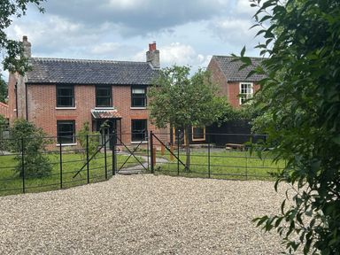 A gravel driveway with a metal gate leading to a brick house with black windows and a tiled roof at Oak Cottage in Somerleyton