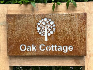 A rusted metal sign with a tree cutout and Oak Cottage text mounted on wood with green leaves at Oak Cottage in Somerleyton
