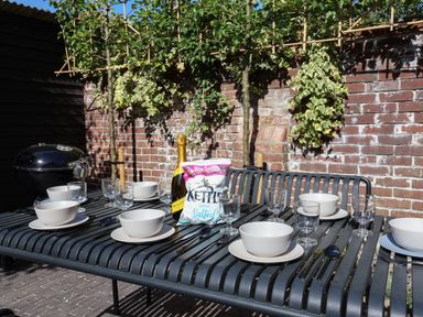 A metal outdoor dining table set with bowls glasses utensils a bottle of wine and a bag of crisps against a brick wall with greenery at Oak Cottage Somerleyton