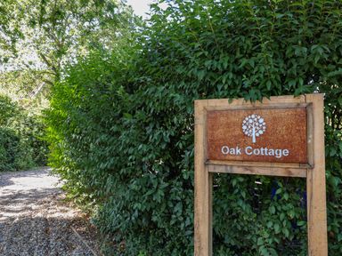 A wooden sign with Oak Cottage text and tree logo next to a gravel pathway and green bushes at Oak Cottage in Somerleyton