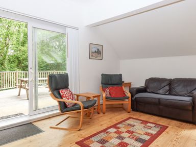 A sitting area with two wooden armchairs with cushions a leather sofa a small wooden table a patterned rug and sliding glass doors leading to a wooden deck at Crystals in Hatherleigh