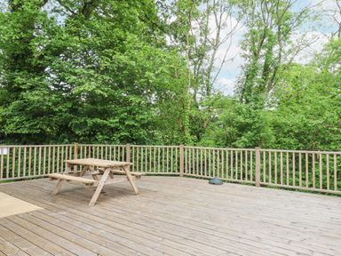 A wooden deck with a wooden picnic table and railing surrounded by trees at Crystals in Hatherleigh