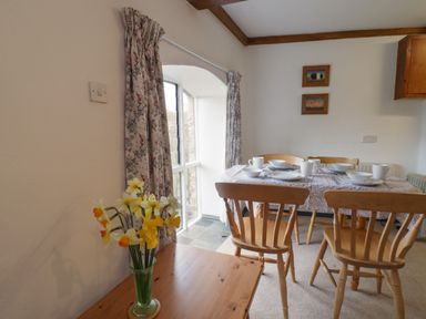 A dining area with a wooden table set with white dishes and mugs four wooden chairs a window with floral curtains and a vase of yellow and white flowers on a wooden side table at Butterton Cottage in Combe Raleigh near Honiton