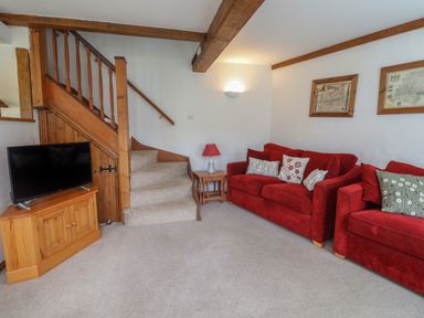 A living room with two red sofas, a wooden staircase, a TV on a wooden cabinet, and framed pictures on the wall at Butterton Cottage in Combe Raleigh near Honiton