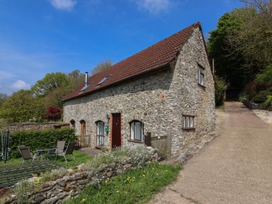 A stone cottage with a tiled roof next to a patio area with chairs and a table at Butterton Cottage in Combe Raleigh near Honiton