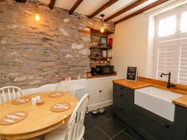 A kitchen with a round wooden table and white chairs next to a stone wall and a sink under a window with shutters at The Nook in Brixham