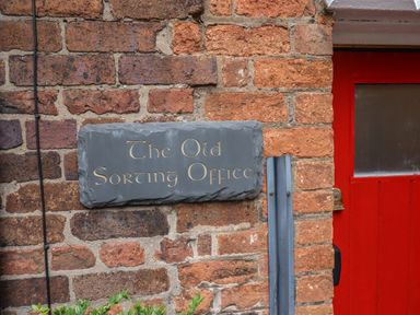 A brick wall with a black slate sign reading The Old Sorting Office next to a red door at The Old Sorting Office in Hawarden