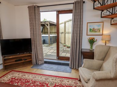 A living room with a TV on a wooden stand a beige armchair a side table with flowers and a lamp and glass doors leading to a patio at The Old Sorting Office in Hawarden