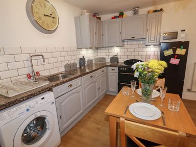 A kitchen with gray cabinets a washing machine a wooden dining table set for two and a black refrigerator at The Old Sorting Office in Hawarden