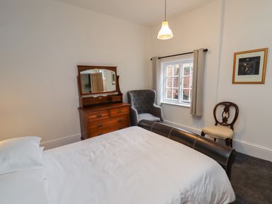 A bedroom with a bed, wooden dresser with mirror, armchair, window, and a wooden chair at The Old Sorting Office in Hawarden