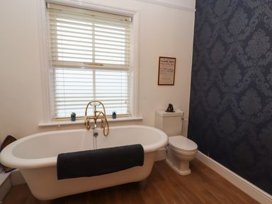 A bathroom with a white bathtub, a towel, a toilet, a window with blinds, and decorative wallpaper at The Old Sorting Office in Hawarden
