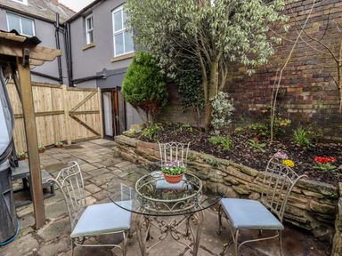 A backyard patio with a glass table and three metal chairs with cushions near a raised garden bed and a wooden fence at The Old Sorting Office in Hawarden