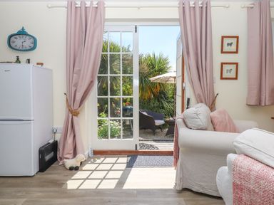 A living area with armchairs and pink curtains by glass doors opening to a garden at Sunset Cottage in Swimbridge near Barnstaple