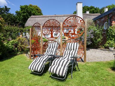 A garden with two striped lounge chairs on grass and decorative metal panels near a thatched roof house at Sunset Cottage in Swimbridge near Barnstaple