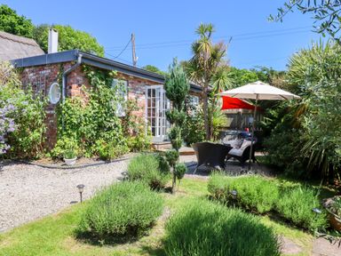 A garden with green bushes and trees surrounding a brick building with an open door and patio furniture at Sunset Cottage in Swimbridge near Barnstaple