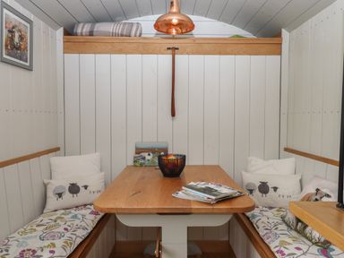A small dining area with a wooden table and cushioned benches with sheep pillows at Burr 1 Shepherds Hut in Barnstaple