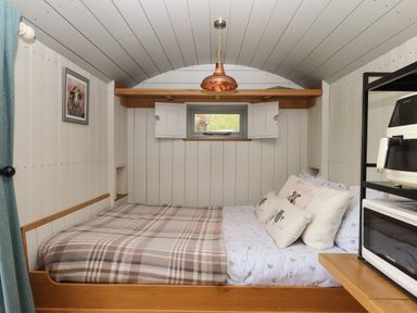 A small bedroom with a bed covered with pillows and a plaid blanket a small window with shutters and a copper ceiling light at Burr 1 Shepherds Hut in Barnstaple