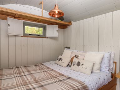 A bedroom with a bed covered with a plaid blanket and pillows under a small window with white shutters at Burr 1 Shepherds Hut in Barnstaple