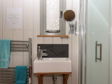 A bathroom with a sink, a small window with frosted glass, a shower with a glass door, a wall-mounted circular mirror, and a towel rack with blue towels at Burr 1 Shepherds Hut in Barnstaple